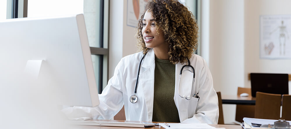 A woman in a white coat sits at a desk, focused on her work with a thoughtful expression.