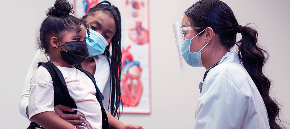 A woman in a white coat and mask gently holds a child, showcasing a caring moment between them.