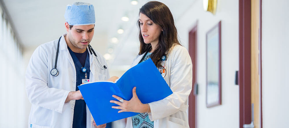 Two physicians talk about patient charts in hospital hallway.