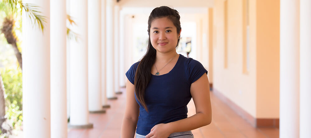 A student wearing a blue shirt stands confidently in a bright hallway, looking ahead with a smile.