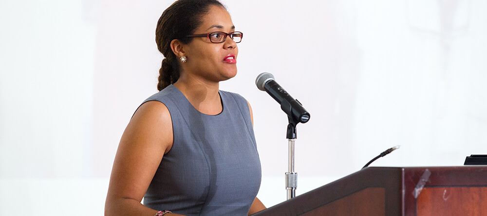 A woman wearing glasses passionately delivers a speech to an engaged audience.